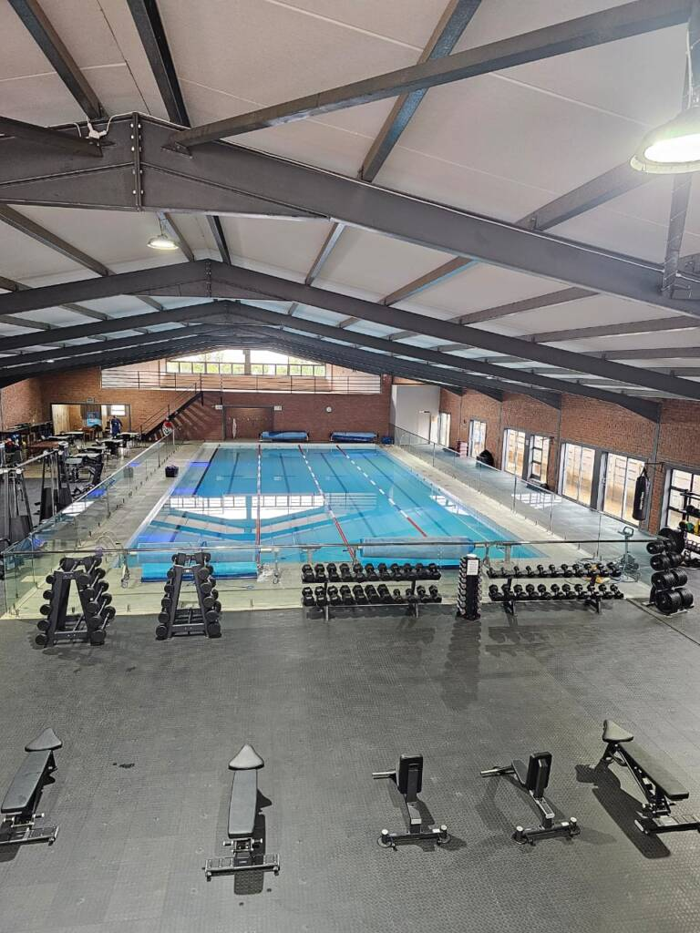 Uitsig oor die JPSS gimnasiumvloer met halterrakke, banke en die binnenshuise swembad agter